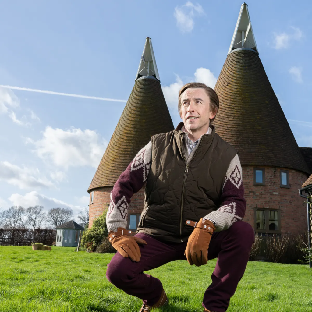 Alan Partridge squatting in front of an oast-style house